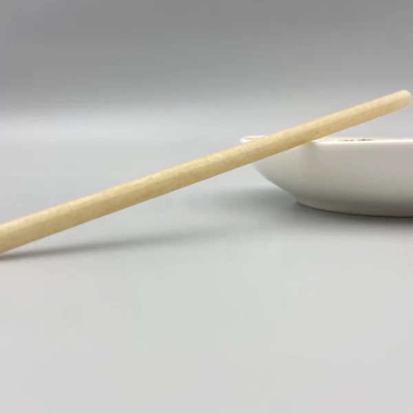 A single sugarcane cocktail straw placed horizontally next to a white ceramic bowl on a neutral gray background